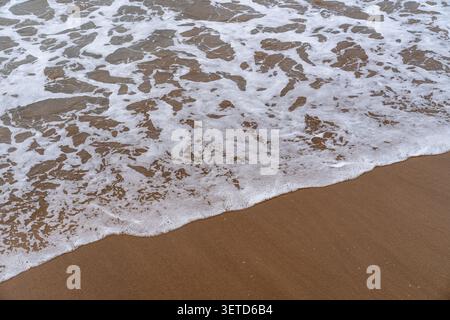De douces vagues océaniques avec de la mousse blanche créent des motifs abstraits lorsqu'elles se jettent sur une plage de sable à Zarautz Banque D'Images