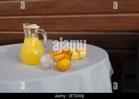 Pichet en verre rempli de limonade jaune pâle, grand verre vide avec des glaçons, citron entier et assiette de tranches de citron disposées sur une table ronde couverte par Banque D'Images