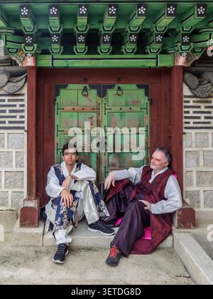 Deux hommes en vêtements masculins Hanbok assis sur des marches devant une porte en bois vert au palais de Gyeongbokgung, Séoul, Corée du Sud. Portrait éditorial. Banque D'Images