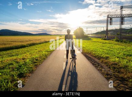 Cycliste féminine avec vélo de route debout sur un chemin asphalté au coucher du soleil Banque D'Images