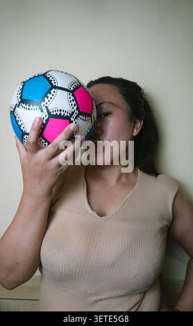 Jeune femme tenant un ballon de soccer devant son visage à l'intérieur, créant un portrait ludique qui représente le sport, les loisirs, le mode de vie actif, et Banque D'Images