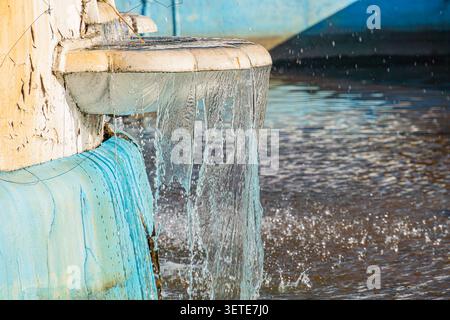 Eau en cascade provenant d'une fontaine en pierre vieillie, avec des ruisseaux clairs, des reflets lumineux de la lumière du soleil, mettant en vedette un tuyau métallique bleu usé, Highligh Banque D'Images