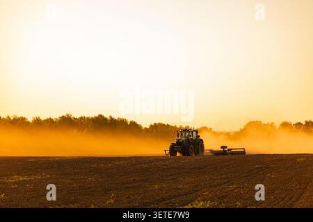 Tracteur travaillant dans le champ avec nuage de poussière au coucher du soleil Golden Hour Banque D'Images