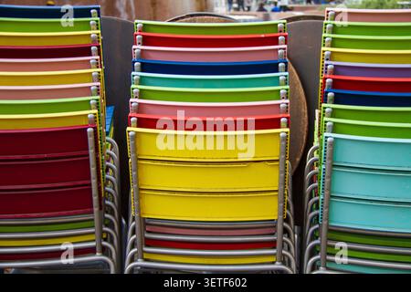 Trois rangées verticales de chaises en métal avec des dossiers en plastique de couleurs vives sont empilées à l'extérieur. Nuances de rouge, jaune, vert, bleu clair et rose A. Banque D'Images