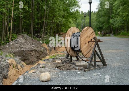 Un grand enrouleur de câble en bois se tient à côté d'une excavation fraîche en bord de route le long d'un chemin de gravier bordé d'arbres verts denses, avec des lampadaires et des travailleurs éloignés à l'intérieur Banque D'Images