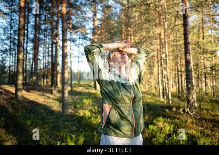 Femme d'âge moyen pratiquant la pleine conscience, se tournant le visage au soleil pendant la promenade dans la forêt de pins sauvage Banque D'Images