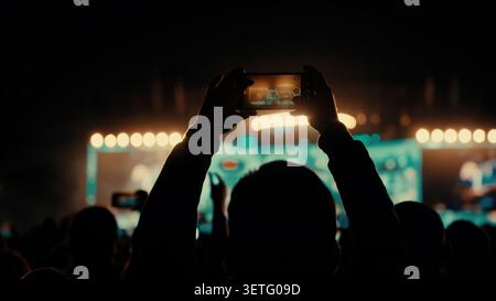 Scène de concert de nuit sombre. Personne silhouette tient un téléphone haut. L'écran affiche les voyants de la scène. Des lumières vives éclairent la scène. La foule forme un Banque D'Images