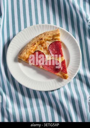 Plan posé à plat au-dessus de la tête d'une délicieuse tranche de pizza au pepperoni salami fraîchement cuite servie sur une assiette en céramique blanche. Le plat repose sur un beauti Banque D'Images