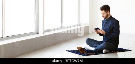 Jeune homme assis sur un tapis de prière lisant un livre religieux à l'intérieur Banque D'Images