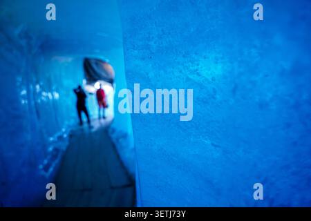 Vue sur les murs de glace bleus vibrants à l'intérieur de la grotte de glace du glacier du Rhône avec les visiteurs marchant le long du chemin en bois à Obergoms, Valais, Suisse. Banque D'Images
