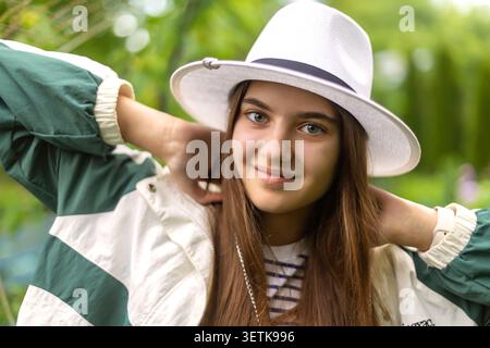 Jeune femme souriante en chapeau blanc à l'extérieur. Jeune femme souriante dans un chapeau blanc et une veste verte pose avec ses mains derrière son cou, entourée par Banque D'Images