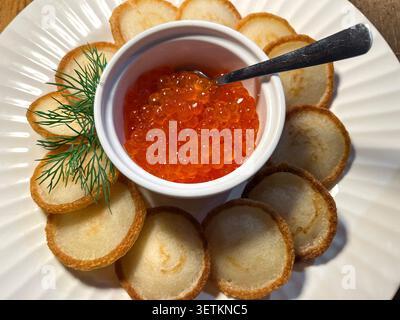 Caviar d'œufs de saumon orange vif servi dans un bol blanc avec une cuillère, entouré de blinis dorés disposés en cercle et d'aneth frais sur une assiette blanche Banque D'Images