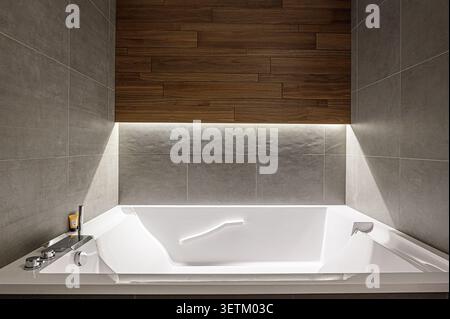La salle de bains moderne dispose d''une baignoire blanche, de murs carrelés gris et de lambris en bois. L'éclairage est doux et améliore le design élégant Banque D'Images
