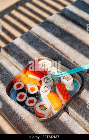 reposant sous des bandes de soleil et d'ombre, un récipient en acier inoxydable rempli de sushis se trouve sur un banc en bois à l'extérieur, avec des baguettes turquoise Banque D'Images