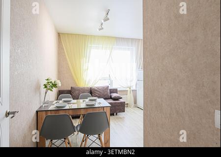 Coin repas confortable avec une table pour quatre. Un canapé confortable se trouve près d'une fenêtre avec des rideaux lumineux. Un vase de fleurs ajoute une touche de nature Banque D'Images