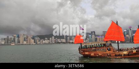 HONG KONG - 18 JUILLET 2023 : la jonque Aqua Luna avec des voiles rouges navigue près de la rive alors que l'horizon de la ville s'illumine au crépuscule. Banque D'Images