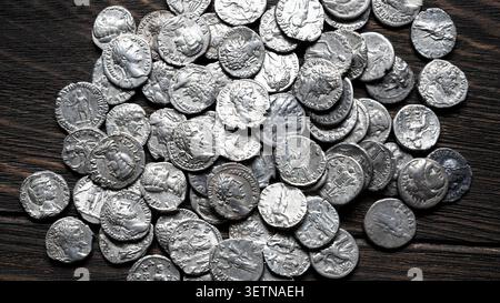 Une photographie en gros plan en grand angle montrant une pile dispersée de pièces de monnaie denarii en argent romain anciennes reposant sur une surface en bois sombre et texturée. Les pièces de monnaie Banque D'Images