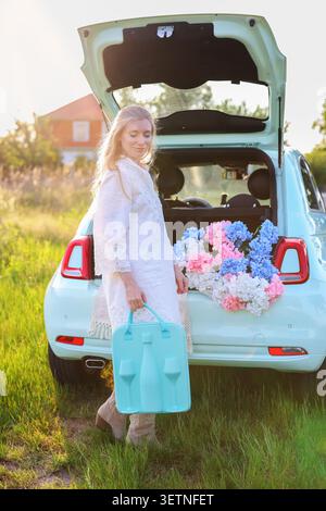 Belle femme blonde profitant de l'heure dorée dans un pré, entourée de fleurs et de lumière douce du soir près d'un véhicule pastel compact Banque D'Images