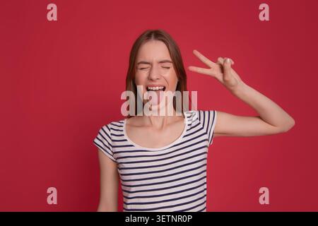 Jeune femme ludique sortant la langue et montrant le signe de paix sur un fond rouge avec espace de copie, plaisir, liberté et concept de style de vie de jeunesse. Banque D'Images