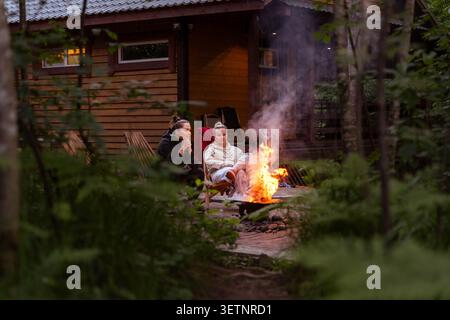 Deux femmes sont assis dans des chaises longues à côté d'un feu de camp incandescent à l'extérieur de la cabane rustique en bois, avec de la fumée qui monte à travers les arbres dans la lumière tamisée du soir Banque D'Images