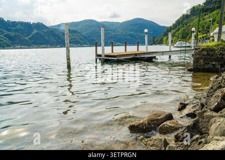 Quai en bois et jetée sur le rivage rocheux du lac de Lugano avec des montagnes vertes en arrière-plan, Caslano, canton du Tessin, Suisse. Jour d'été nuageux. NAT Banque D'Images