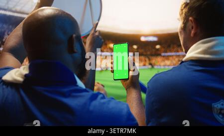 Chroma Key smartphone utilisé par l'homme dans le stade de football se tient pour suivre les scores en direct. Un supporter de football afro-américain vérifie l'application de Paris sur le téléphone à écran vert pendant le jour du match Banque D'Images
