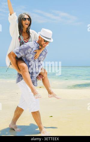 Jeune femme joyeuse jouant à sauter s'appuyant sur des hommes de retour le long de la plage de sable turquoise du rivage de l'océan plage de sable globale île tropicale destination de villégiature ensoleillée Banque D'Images