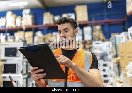 Jeune homme portant un gilet de sécurité exploitant une tablette numérique dans un centre de distribution, gérant diligemment le stock et assurant un fonctionnement efficace de l'entrepôt Banque D'Images
