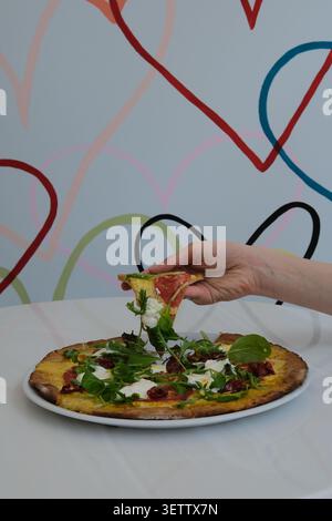 Main soulevant la tranche de pizza de l'assiette avec des légumes verts, du fromage, de la tomate, un repas rustique décontracté Banque D'Images