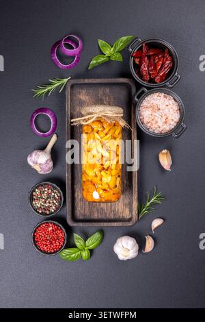 Champignons au miel marinés dans un bocal en verre centralisé sur un plateau en bois rustique, entouré d'ail, d'oignon rouge, de basilic, de romarin, divers grains de poivre, dri Banque D'Images
