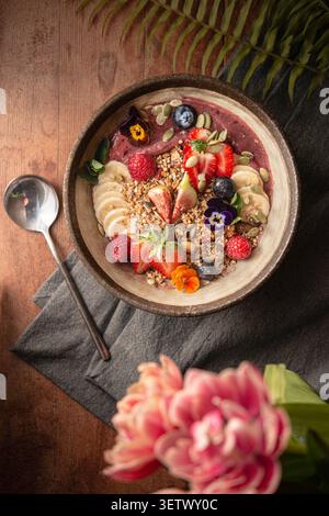 Bol à smoothie à l'açai vibrant surmonté de fraises fraîches, figues, bananes, granola et fleurs comestibles. Banque D'Images