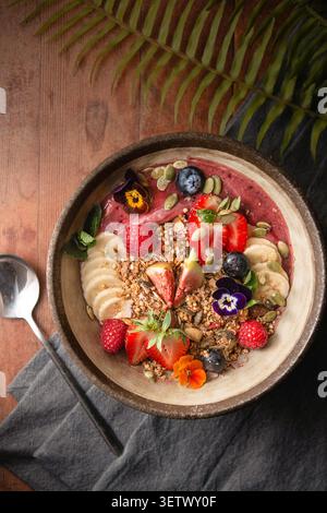 Bol à smoothie à l'açai vibrant surmonté de fraises fraîches, figues, bananes, granola et fleurs comestibles. Banque D'Images