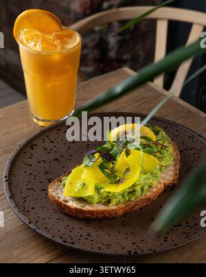Pain grillé à l'avocat au levain garni de légumes jaunes marinés et de microverts, servi avec un verre de jus d'orange glacé. Banque D'Images