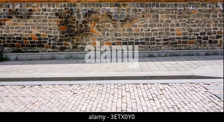 Vieux mur de briques avec pierres altérées et mortier. La chaussée pavée s'étend vers le mur. Un rebord en béton sépare le mur du sol Banque D'Images