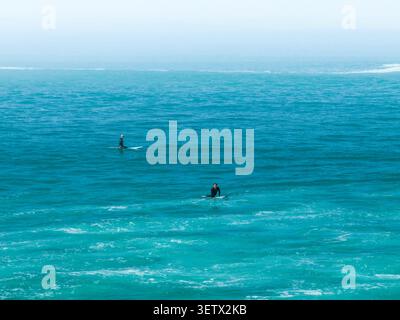 Deux surfeurs attendent sur leurs planches dans une eau turquoise calme, vue d'en haut contre un horizon brumeux. Banque D'Images