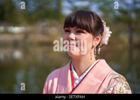 Portrait en gros plan d'une jeune femme adulte dans un kimono rose avec accessoire de cheveux floral et boucles d'oreilles pendantes, regardant avec une expression sereine à côté d'un étang Banque D'Images