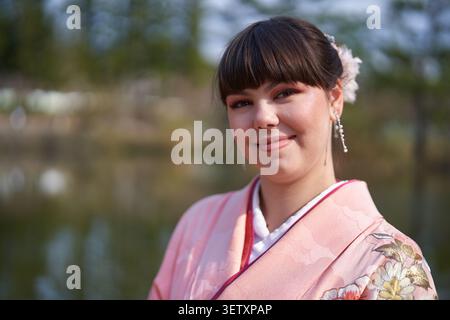 Portrait en gros plan d'une jeune femme adulte dans un kimono rose avec accessoire de cheveux floral et boucles d'oreilles pendantes, regardant avec une expression sereine à côté d'un étang Banque D'Images
