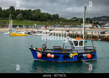 Un petit bateau de pêche bleu flotte dans le port de Lyme Regis avec des bouées colorées sur le côté et une plage de sable et une ville en arrière-plan Banque D'Images