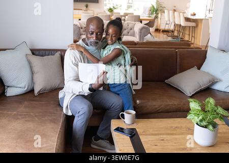 Homme afro-américain et jeune fille assis sur un canapé en cuir, tenant du papier et montrant le sac à dos Banque D'Images