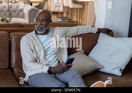Homme afro-américain senior assis sur un canapé sectionnel à la maison, tenant un smartphone, portant un écouteur Banque D'Images