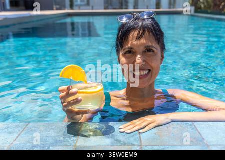 Femme mature en maillot de bain bleu lunettes de soleil debout dans la piscine carrelée tenant cocktail avec tranche d'orange Banque D'Images