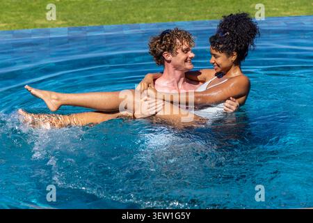 Couple adulte se tenant dans la piscine de l'arrière-cour, éclaboussures d'eau, maillot de bain blanc une pièce, ombres Banque D'Images