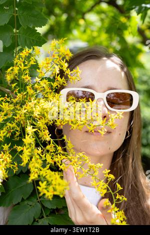 Jeune femme portant des lunettes de soleil blanches parmi les fleurs sauvages jaunes dans le jardin d'été Banque D'Images