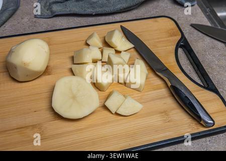 Pommes de terre fraîchement pelées coupées en morceaux sur une planche à découper en bois avec un couteau de cuisine. Cuisine maison, préparation des repas et concept de préparation des aliments. Banque D'Images