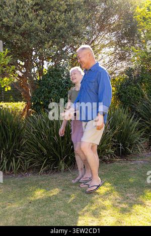 Couple senior diversifié marchant main dans la main sur la pelouse du jardin herbeux, portant des tongs et montre-bracelet Banque D'Images