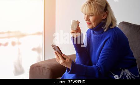 Femme senior se relaxant sur un canapé confortable, s'engageant joyeusement avec son smartphone tout en profitant d'une tasse de café chaud à l'intérieur près d'une fenêtre lumineuse Banque D'Images