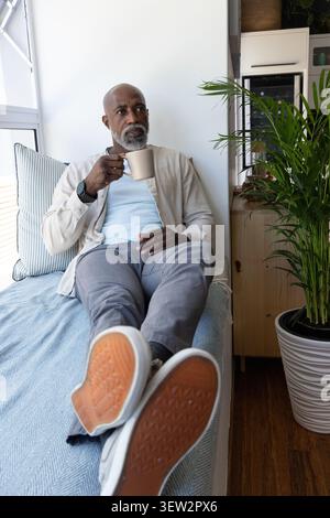 Homme africain senior s'inclinant sur un lit de jour près d'une grande fenêtre, tenant une tasse en céramique, portant un tee-shirt bleu clair Banque D'Images
