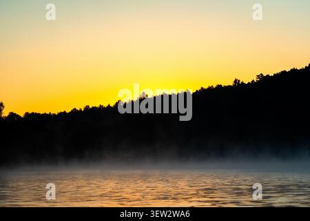 Un ruban pâle de soleil borde la crête tandis que la brume glisse sur la surface de la rivière. Les petites vagues attrapent des couleurs douces et la vallée s'illumine lentement dans un calme Banque D'Images