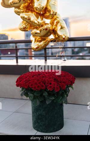 Cadre romantique de célébration surprise sur le balcon du toit avec un énorme bouquet de roses rouges de luxe dans une boîte et un paquet de ballons de coeur en feuille d'or. Blurre Banque D'Images