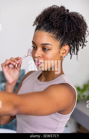 Femme afro-américaine adulte utilisant le rouleau de quartz rose sur la joue à la maison portant haut, cerceaux en or Banque D'Images